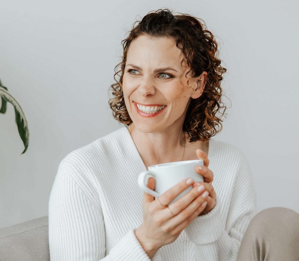 Andrea sitzt lachend auf der Coach mit einer Kaffee Tasse in der Hand.