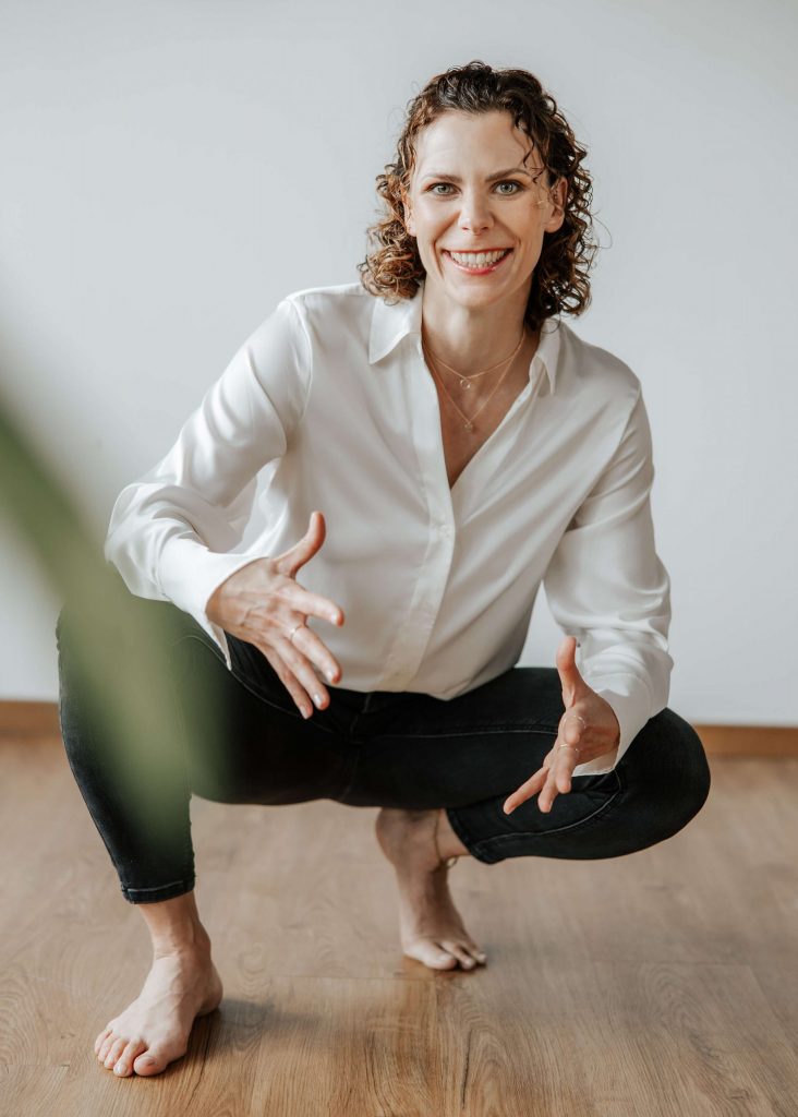 Andrea Görlich in weißer Bluse und dunkler Hose, barfuß in der Hocke auf einem Holzfußboden, mit offener, einladender Geste und Lächeln.