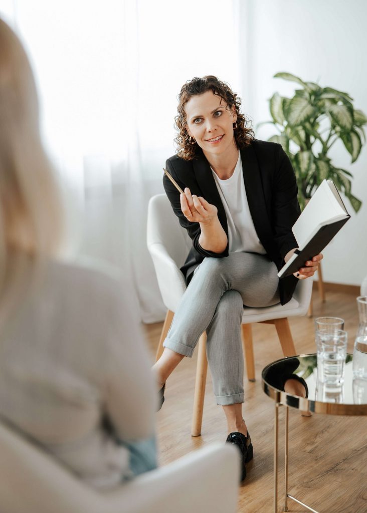 Andrea Görlich sitzt lächelnd auf einem Stuhl, zeigt ein offenes Notizbuch und spricht mit einer Klientin, Blickkontakt und professionelle Atmosphäre.