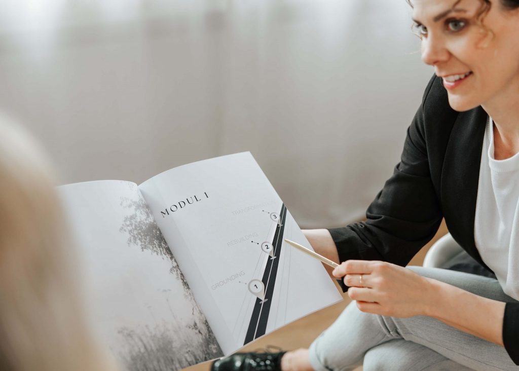 Nahaufnahme eines geöffneten Workbooks mit Modulübersicht, Andrea zeigt mit einem Stift auf die Inhalte – strukturiertes Coachingmaterial.