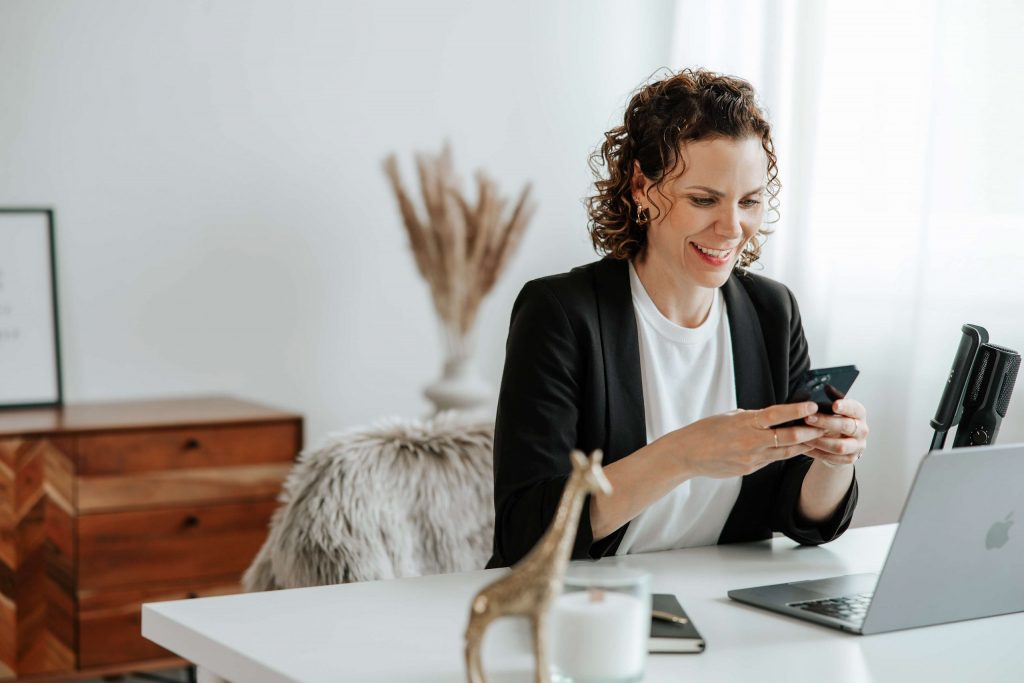 Andrea Görlich sitzt an einem hellen Schreibtisch, lächelt beim Blick aufs Handy, Laptop und Mikrofon im Vordergrund – digitale Coaching-Szene.