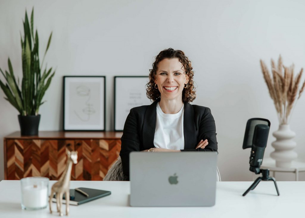 Frau sitzt lächelnd am Schreibtisch mit Laptop, Mikrofon und Deko, beide Arme vor der Brust verschränkt.