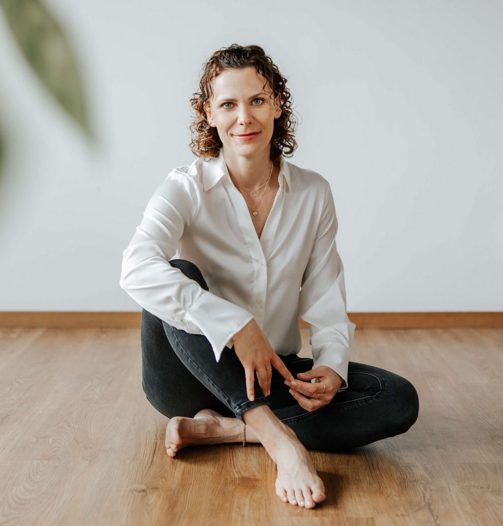 Andrea Görlich sitzt mit überkreuzten Beinen auf dem Boden und schaut ruhig in die Kamera, eine Pflanze ist im Vordergrund unscharf zu sehen.