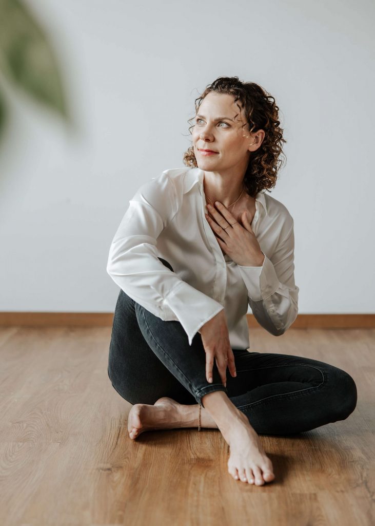 Andrea Görlich blickt leicht zur Seite, eine Hand an der Brust, sitzend auf dem Boden mit natürlicher Ausstrahlung.